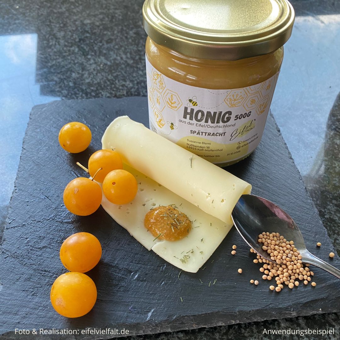 gelbe Pflaumen und Honig und Senfkörner auf einer Schieferplatte und einem Klecks Pflaumensenf auf dem Käse