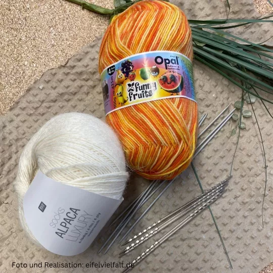 Material: Frunny Fruits Sockenwolle von Opal in orange, Alpaca Luxury Socks in weiß und 2 Nadelspiele in 2,5mm und 1 Kone Make-it-Blümchen in Efeu und eine passende Stricknadel liegen auf einem weißen Hintergrund