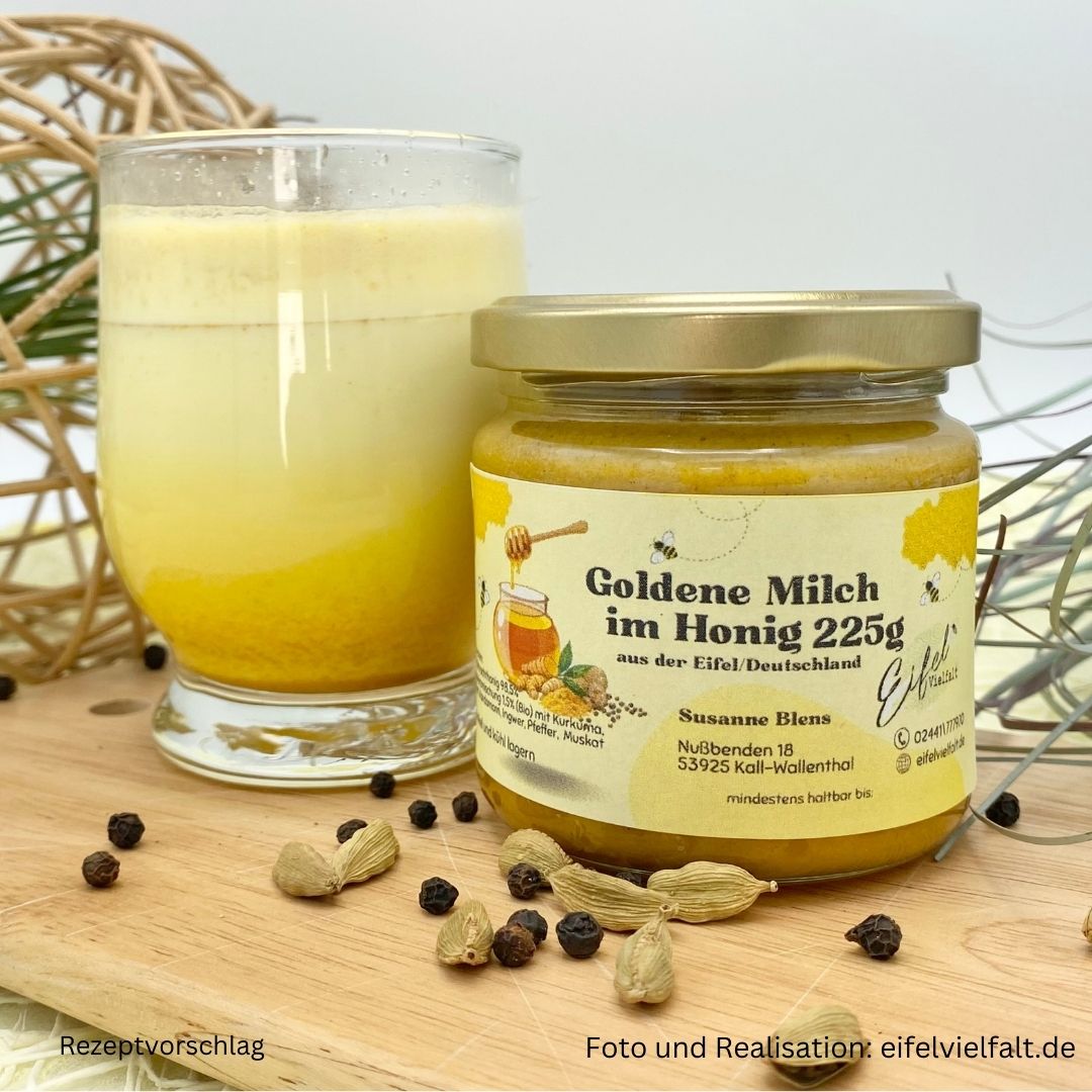 Goldene Milch im Honig im 225g Glas auf Holzbrett dekoriert mit Kardamom und schwarzem Pfeffer und einer zubereiteten MIlch von vorne fotografiert